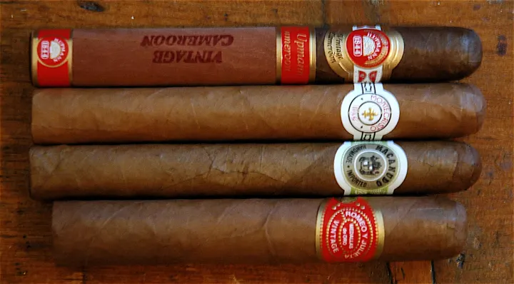 Four cigars stacked on a wooden table, wrappers ranging from mid brown to dark brown.
