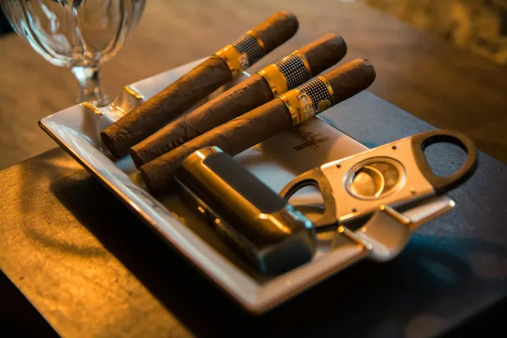 A double-blade guillotine cutter on an ashtray next to three cigars and a lighter.