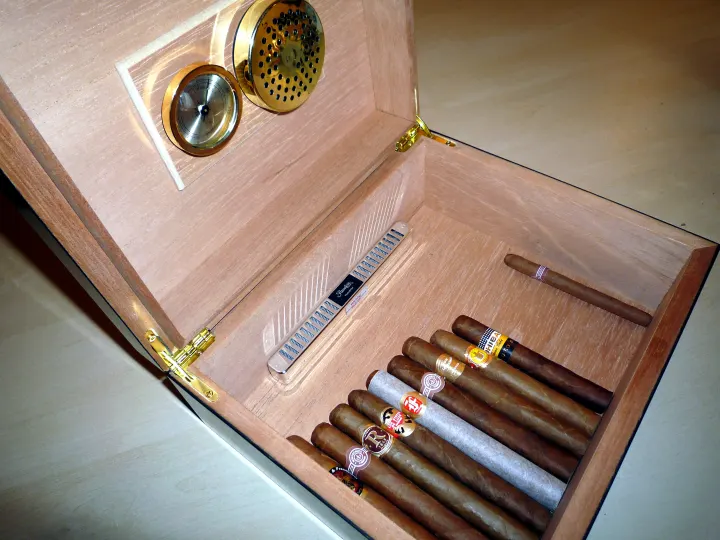 An open wooden humidor with a round hygrometer and a humidifier in the lid, and cigars inside.