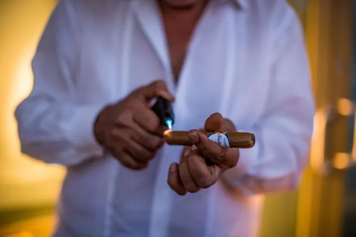 A man toasting the foot of a cigar with a blue-flame butane torch lighter.